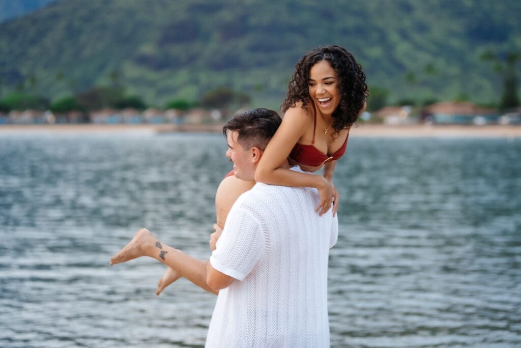 A man lifts a woman in a white dress while embracing near the ocean with mountains in the background during daytime.
