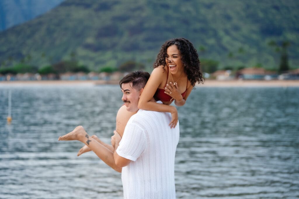 A woman in a white dress holds a young child while standing in shallow ocean water with a sandy beach and mountains visible in the background.