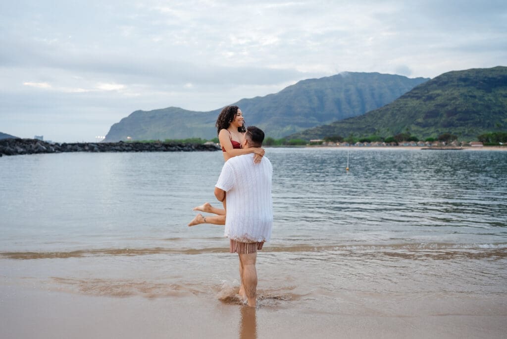 A young child in a white dress stands in shallow ocean water on a sandy beach with dramatic mountainous coastline in the background.