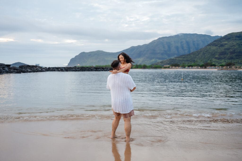 A woman in a white dress stands at the water's edge on a beach with mountains visible in the background under a cloudy sky.