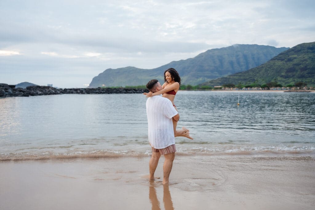 A woman in a white dress stands in shallow beach water with a scenic mountainous coastline in the background.