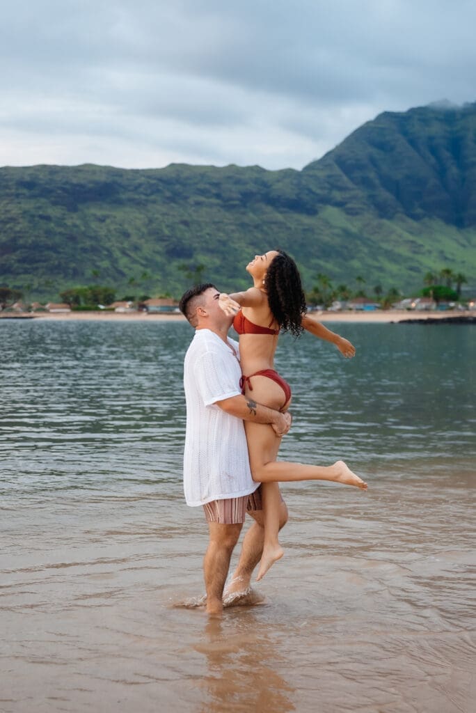 A couple enjoys a romantic moment in shallow ocean water with tropical mountains in the background on a cloudy day.