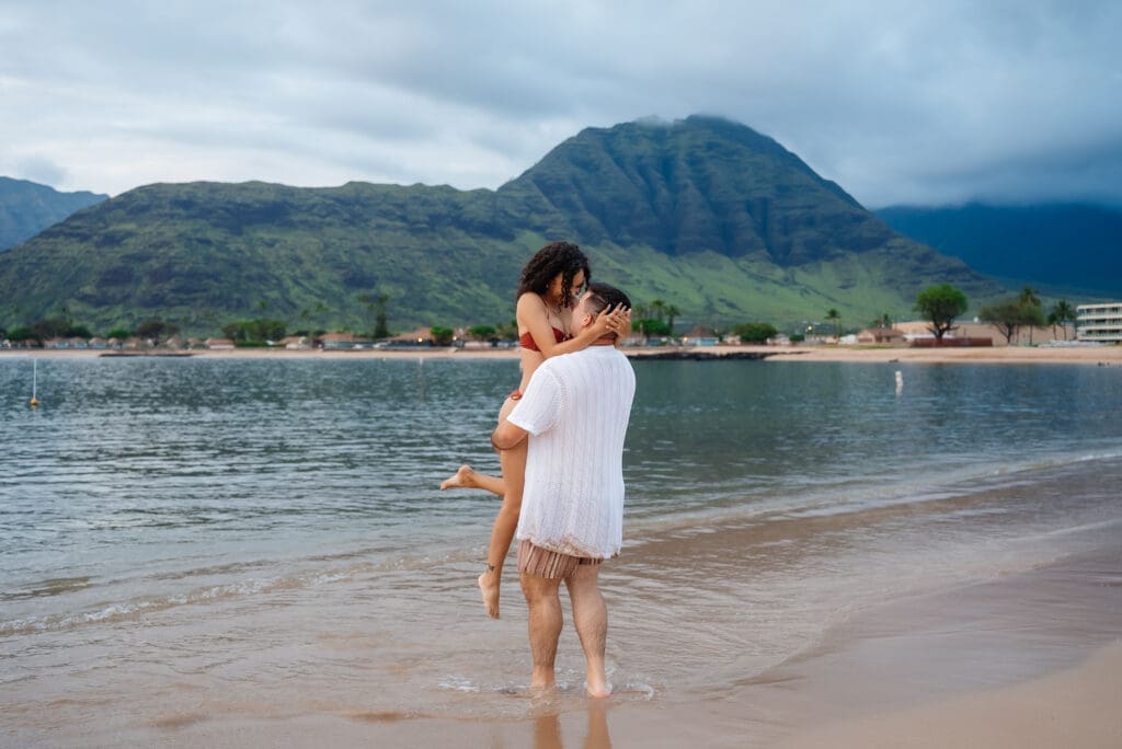 A couple embraces while standing in shallow ocean waters on a tropical beach with mountains visible in the background.