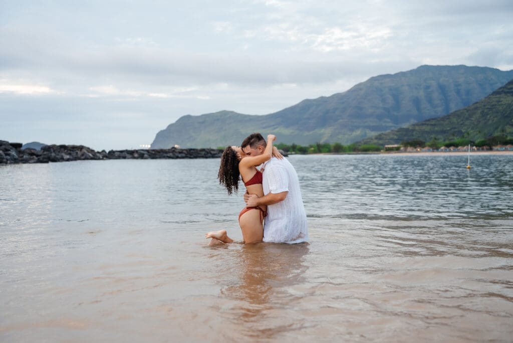 A couple shares a romantic moment in shallow ocean water during their beach wedding ceremony with mountains visible in the background.