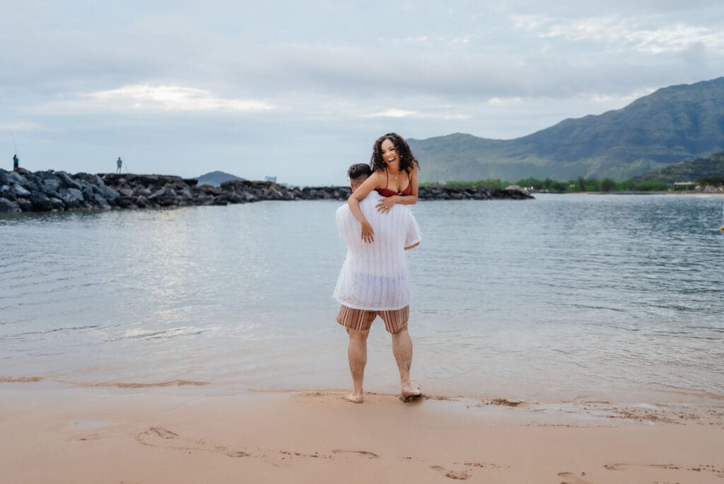 A woman in a white dress stands waist-deep in calm ocean water at a scenic beach with mountains and a rocky pier visible in the background.