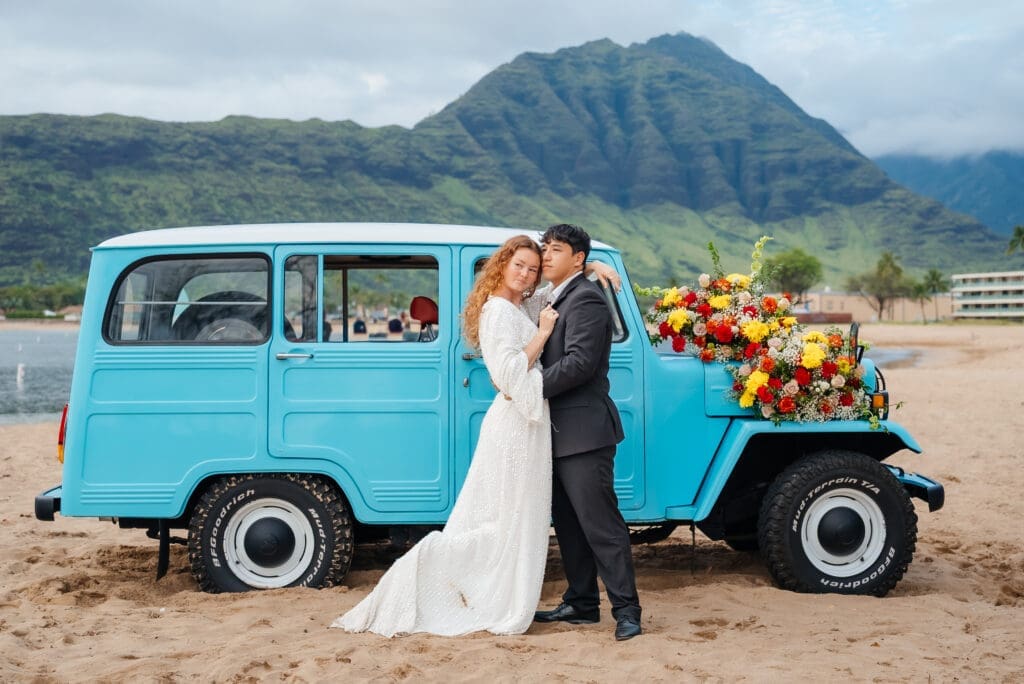 A bride and groom pose together against a vintage turquoise truck decorated with colorful flowers on a scenic beach with mountains in the background.