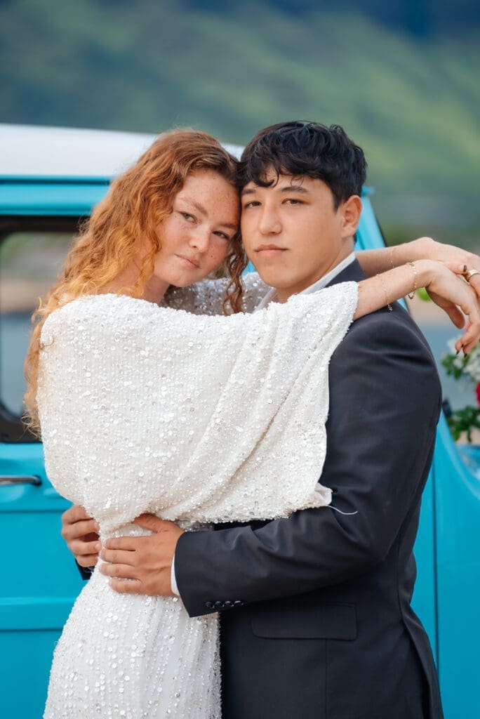 A couple in formal wedding attire poses together against a blue vintage vehicle with a scenic outdoor backdrop.