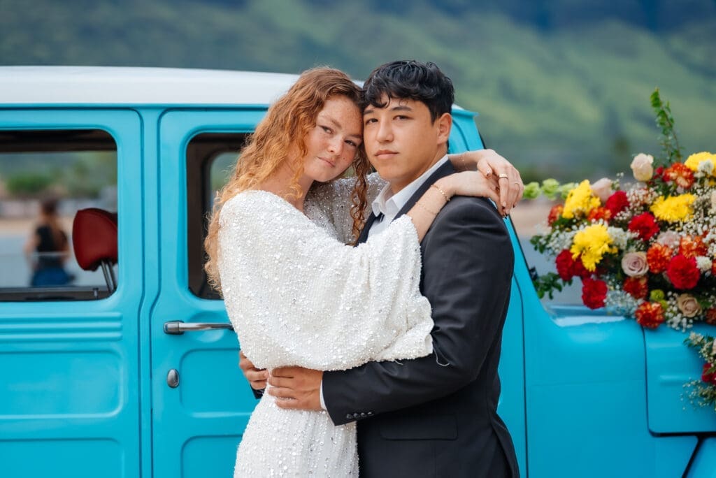 A bride and groom embrace in front of a vintage turquoise truck decorated with colorful flowers in a mountain setting during golden hour.