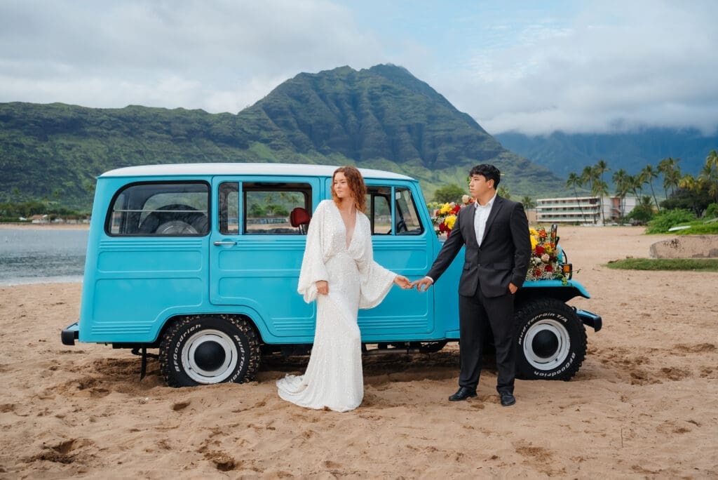 A bride and groom pose together beside a vintage turquoise van on a beach with mountains in the background during their wedding day.