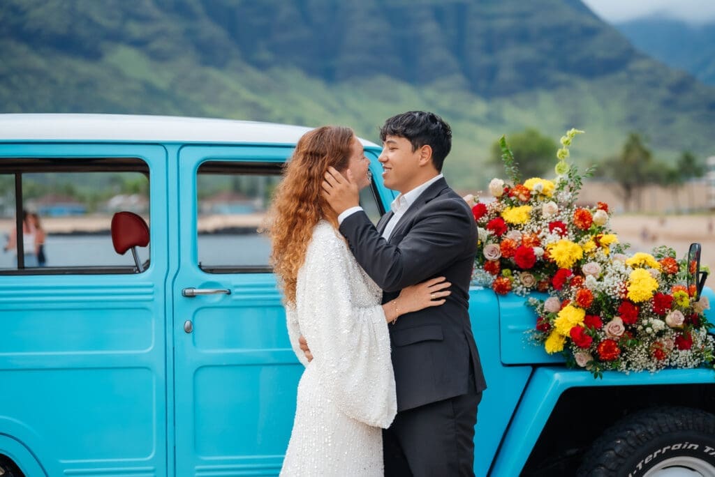 A bride and groom pose together beside a bright turquoise vintage truck decorated with colorful flowers in a mountain valley setting.