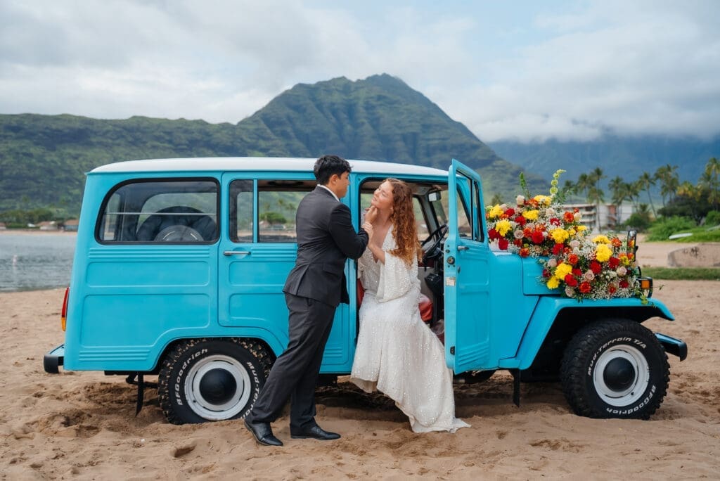 A bride and groom pose beside a bright turquoise vintage van decorated with flowers on a beach with mountains in the background.