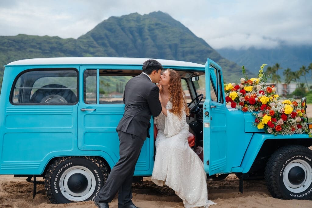 A bride and groom share a romantic moment leaning against a vintage turquoise van decorated with colorful flowers, with a majestic mountain backdrop.