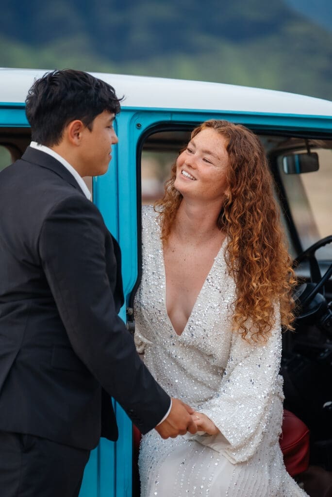 A bride and groom pose together in front of a blue vintage vehicle, appearing to be at a wedding celebration.