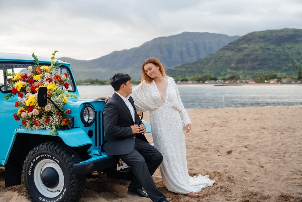 A bride and groom pose together next to a vintage turquoise truck decorated with flowers on a scenic beach with mountains in the background.