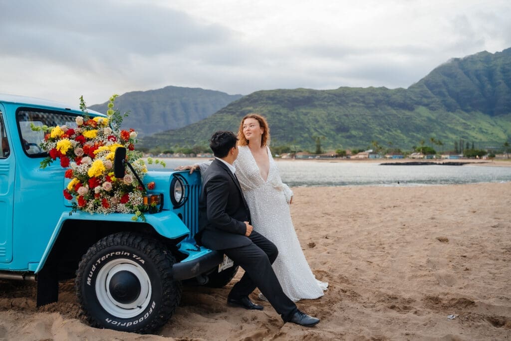 A bride in a white dress poses against a turquoise vintage truck decorated with yellow flowers on a beach with mountains in the background.