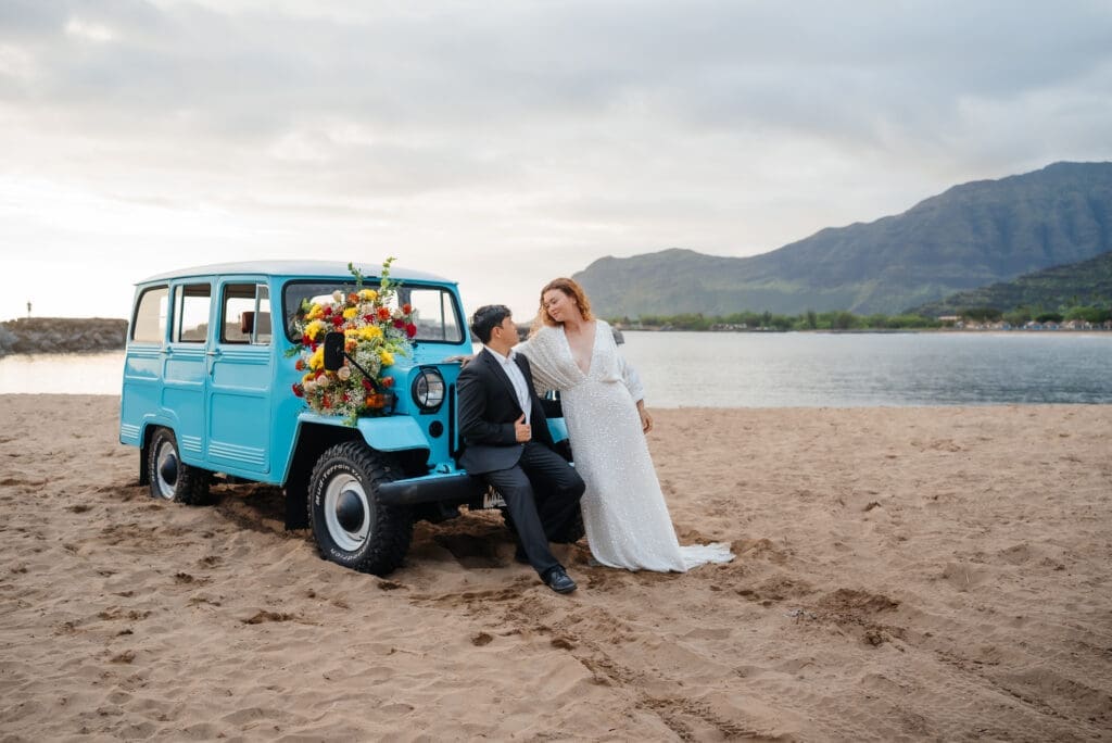 A bride and groom pose together next to a vintage turquoise van on a sandy beach with mountains in the background.