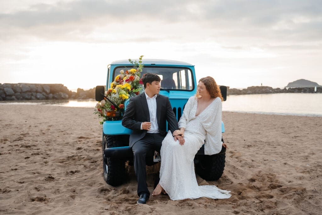 A bride and groom pose with a blue vintage truck on a sandy beach during their wedding day.