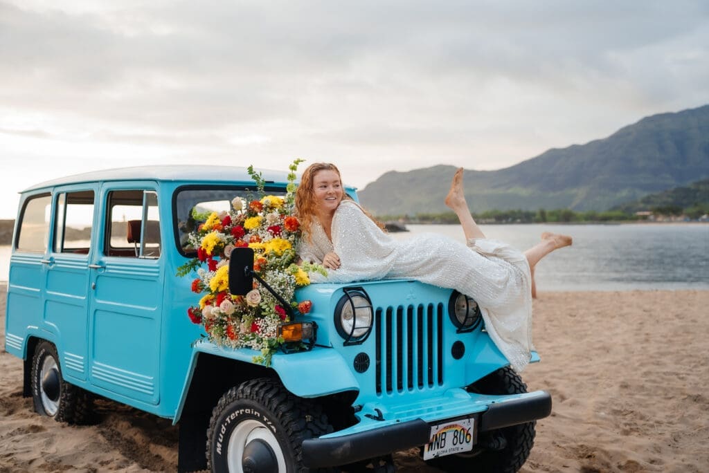 A woman in a white dress poses with a turquoise vintage Jeep decorated with sunflowers on a beach at golden hour, with mountains visible in the background.