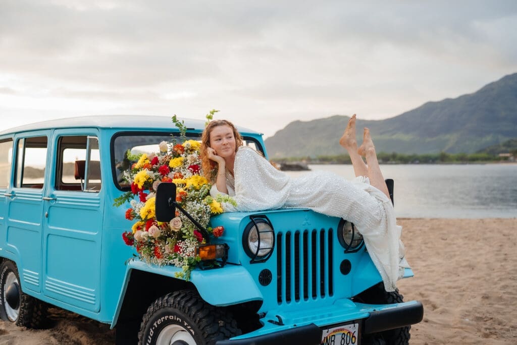 A woman poses with a large sunflower bouquet against a vintage turquoise Jeep at a scenic lakeside location with mountains in the background.