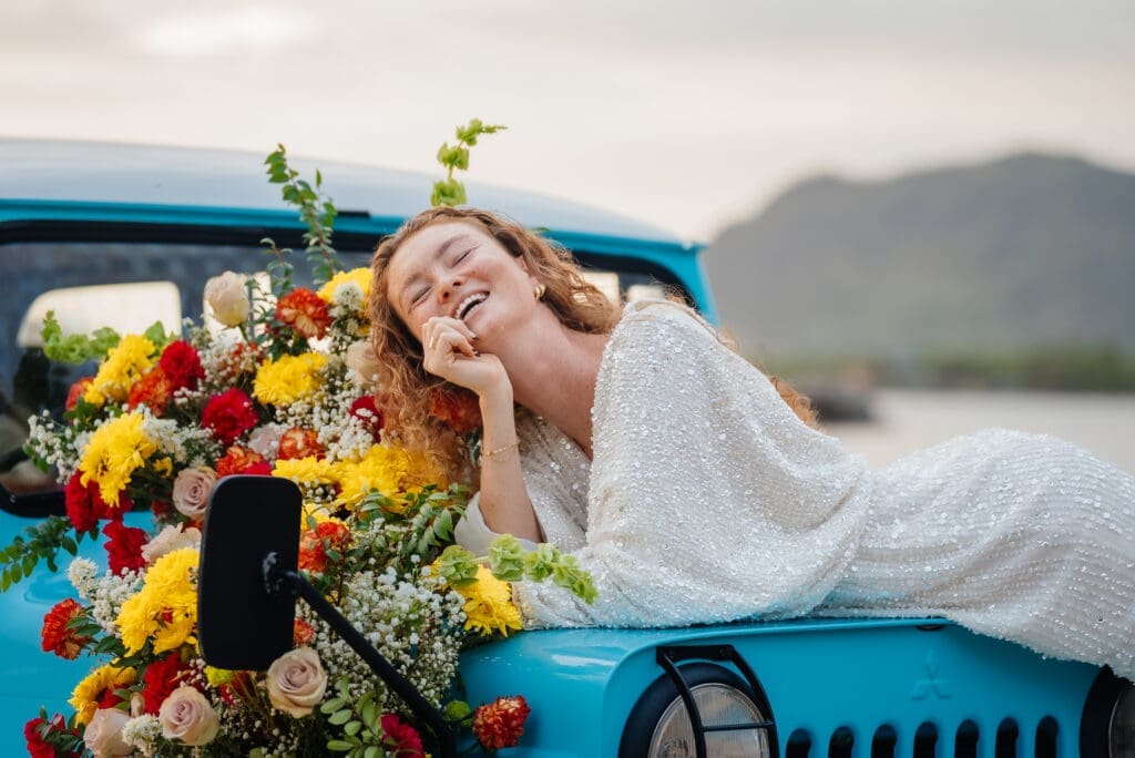 A joyful bride in a white wedding dress reclines on a vintage turquoise truck bed adorned with vibrant colorful flowers, photographed during golden hour with mountains and water in the background.