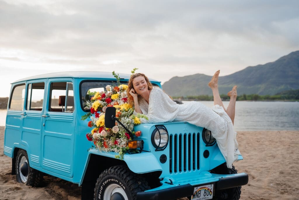 A bride and groom pose joyfully with a turquoise vintage jeep decorated with flowers on a beach, with mountains visible in the background.