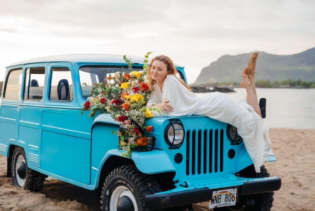 A bride in a white dress poses on a vintage turquoise Jeep decorated with colorful flowers on a beach with mountains in the background.