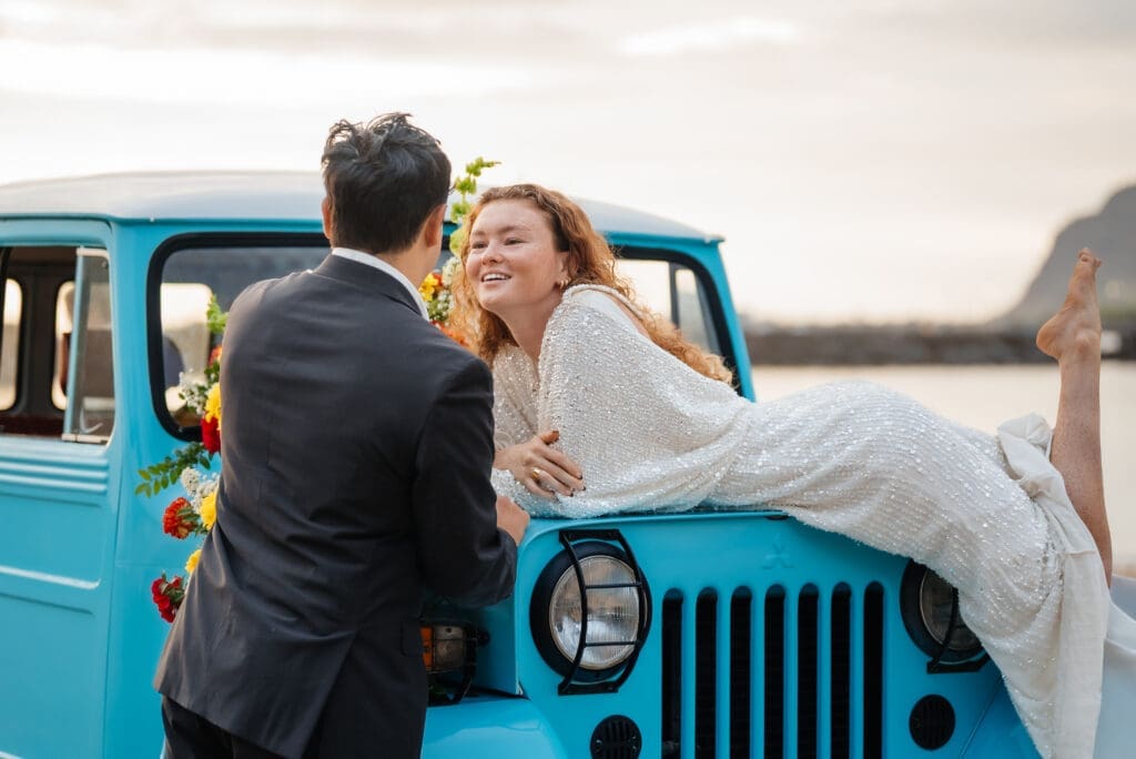 A bride and groom share an intimate moment on a vintage turquoise jeep decorated with flowers during their wedding celebration at golden hour.