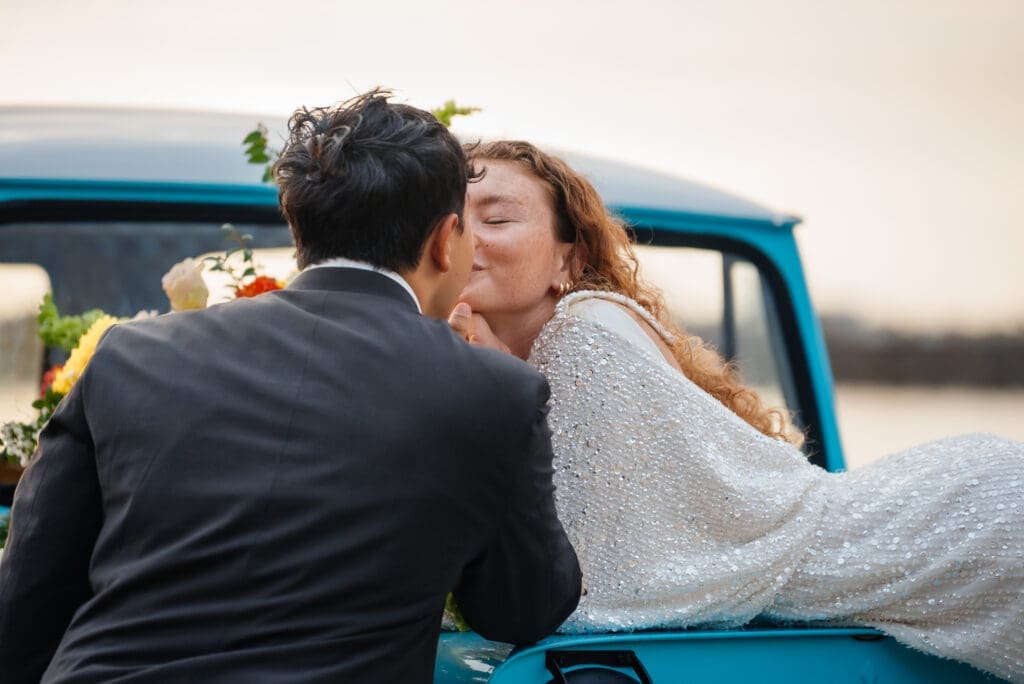 A bride and groom kiss beside a vintage blue truck during golden hour, with flowers visible in the background.