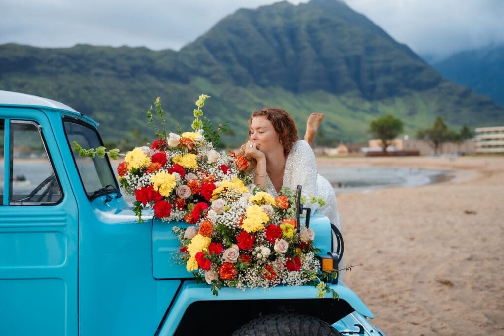 A woman poses with a vibrant bouquet of red and yellow flowers displayed in the bed of a turquoise vintage truck on a scenic beach with mountains in the background.