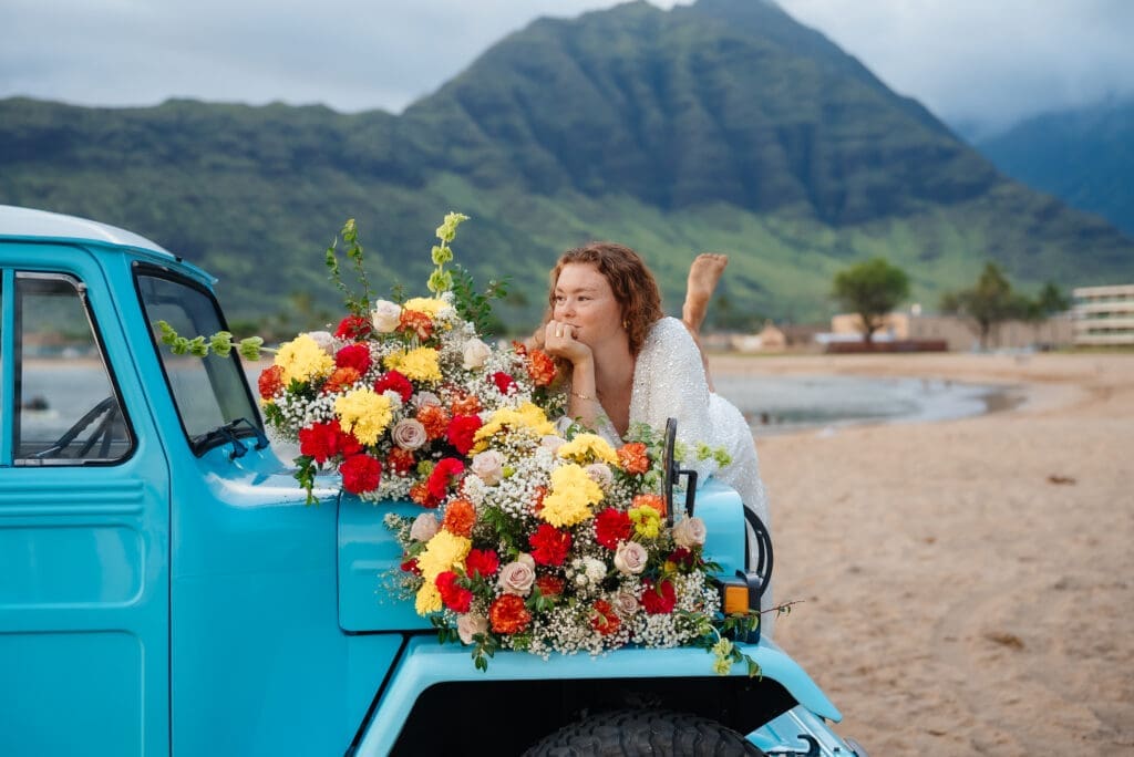 A woman poses with a vibrant bouquet of red and yellow flowers on a vintage turquoise truck bed against a mountainous coastal landscape.