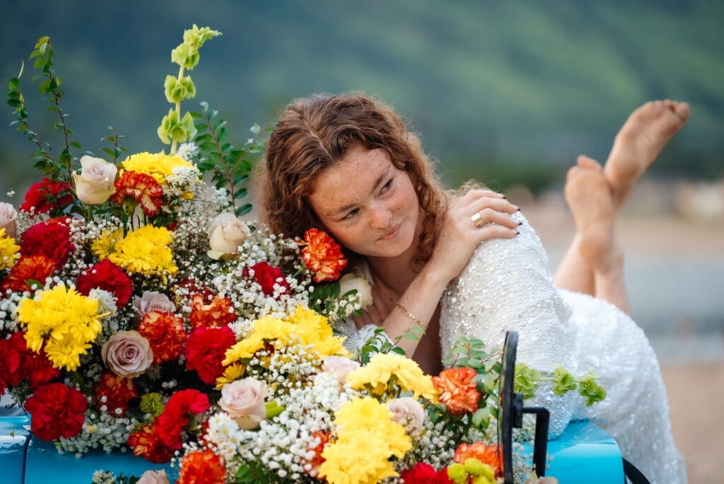 A smiling woman in a white shirt poses with a large vibrant bouquet of red, yellow, and white flowers against a blue water backdrop during daytime.